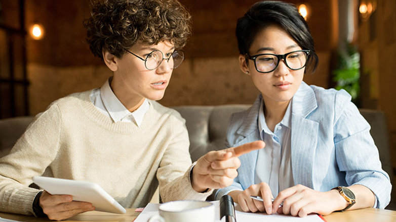 Two people are sitting at a table having a meeting. One has short brown hair and is wearing glasses, a white button-down shirt, and a tan sweater. She is holding a tablet and pointing at it while she talks. The other person has long dark hair and is wearing glasses, a blue blazer, and a white button-down shirt. She is looking at the tablet.