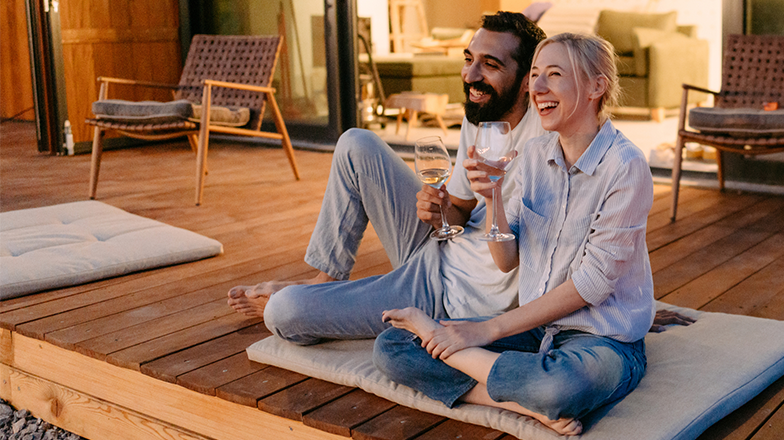 A man and woman sit facing forward with big smiles on a wooden deck, each holding a glass of white wine. They are angled towards each other, and the woman's bare foot is touching the man's calf. The evening sun is casting a warm glow on them.