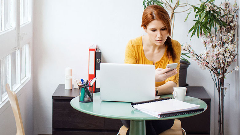 Woman sitting at her desk with her laptop and mobile phone in her hand.