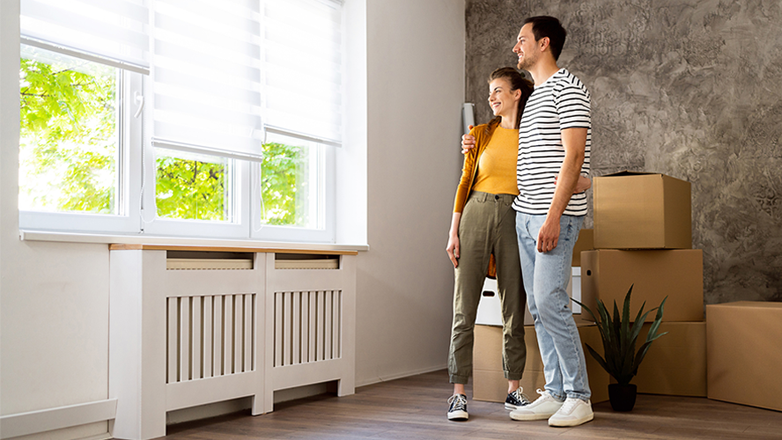 A young couple is standing in a new house, looking out the window. The woman is smiling and the man has his arm around her shoulder. There are boxes and a plant next to them.