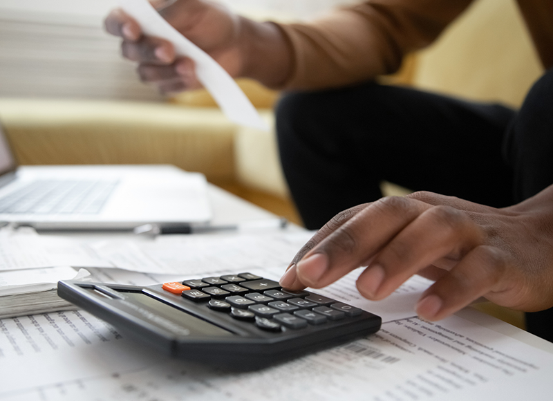 A person is sitting at a desk and using a calculator to add up numbers on a piece of paper. There is an open laptop on the desk.