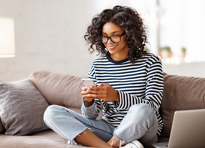 Young woman uses online banking on her phone while relaxing on her sofa.