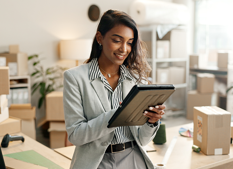 A young woman stands in an office with boxes stacked behind her. She looks down at a tablet, checking details and smiling. The background is out of focus, showing a cluttered office space with more boxes, a potted plant, and a desk with a computer. The woman's smile suggests she is confident and content with her work.