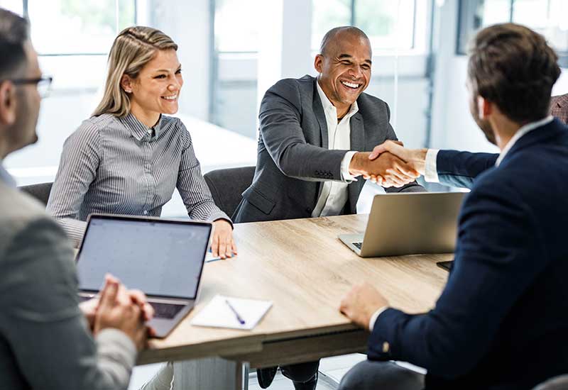 Four people sit at a table, two men are shaking hands while two other people, a woman and a man, look on. There are two laptops on the table, along with a pen and notepad.