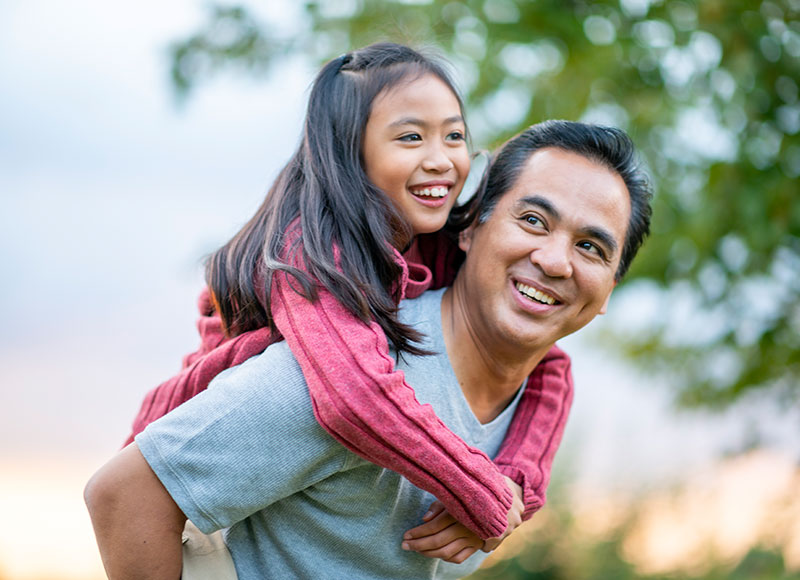 A father gives his young daughter a piggyback ride outside on a sunny day.